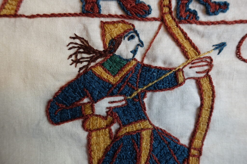 An embroidered Norman archer, hair flying, pulling the arrow back to discharge it. 